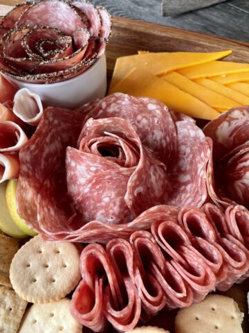 5 different salami roses on a charcuterie board with crackers sliced apples and cheese triangles