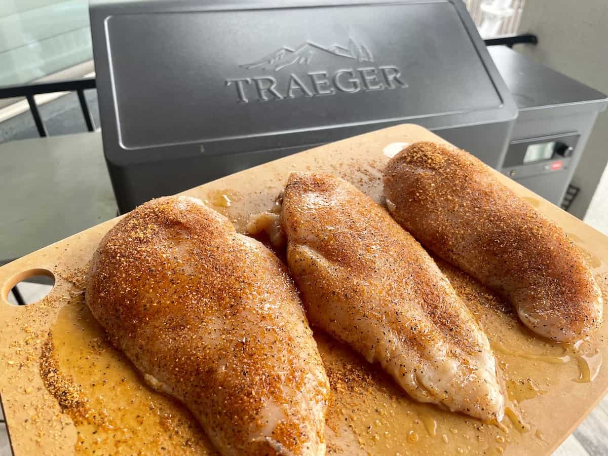 3 seasoned chicken breasts on cutting board in front of traeger grill