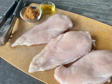 three raw chicken breasts on a wood cutting board with a bowl of rub and bowl of olive oil