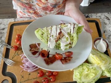 assembled wedge salad on a white plate held up over a cutting board of wedge salad ingredients