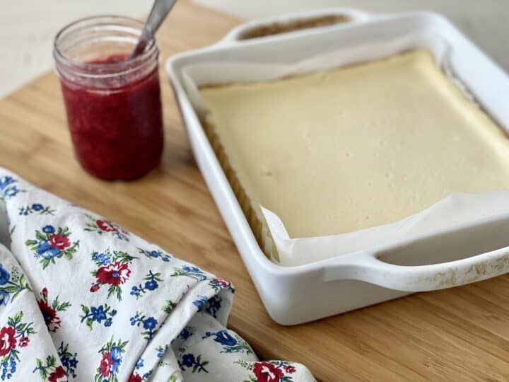 baked cheesecake in white dish next to jar of strawberry sauce and floral apron