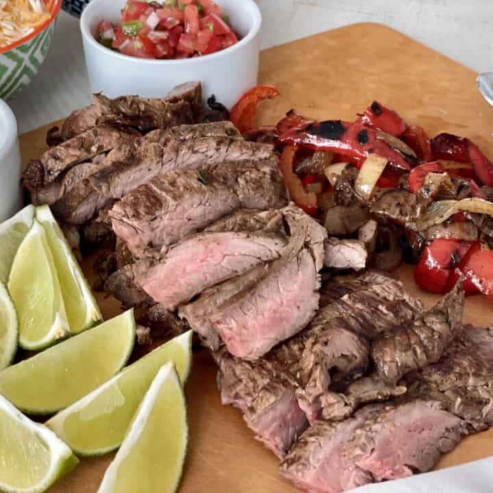 sliced fajita steak on a cutting board with lime wedges and cooked bell pepper and onions