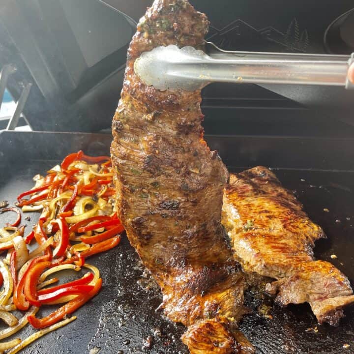 skirt steak held up with tongs over Traeger flatrock with peppers and onions cooking