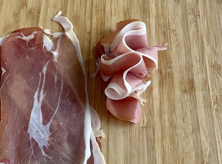 full slice of prosciutto and prosciutto ribbon in a wood cutting board