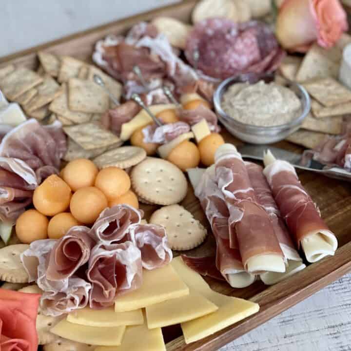 large wood board showcasing prosciutto folded in several different ways with crackers cheese and mustard