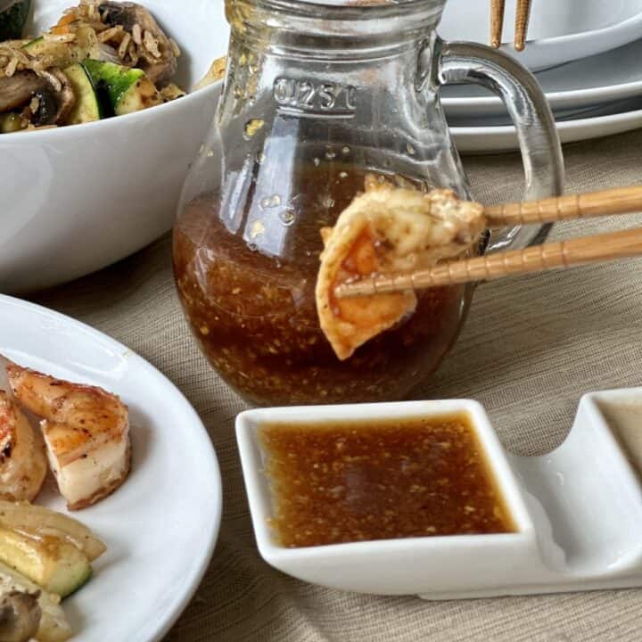 hibachi shrimp in chopsticks with white dish of ginger dipping sauce and glass pitcher of sauce