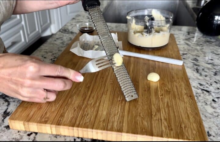 hand hold fork piercing garlic clove while grating on microplane over wood cutting board with mini food processor in background 