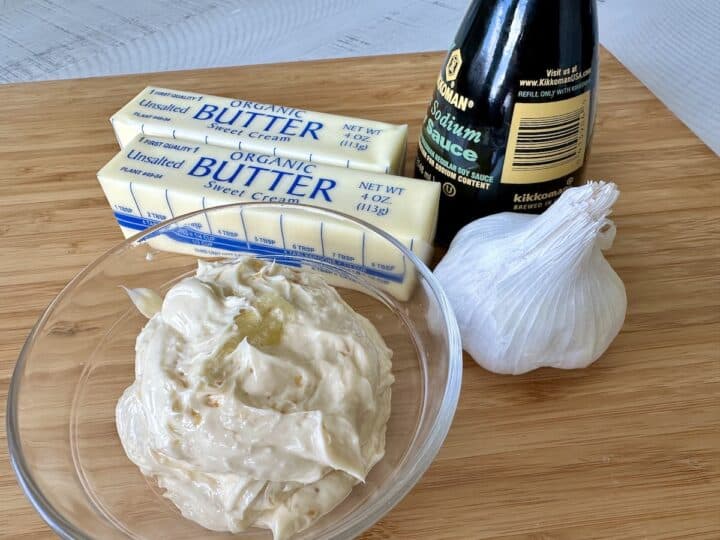 prepared hibachi garlic butter in a glass bowl next to 2 sticks of butter head of garlic and soy sauce on wood cutting board