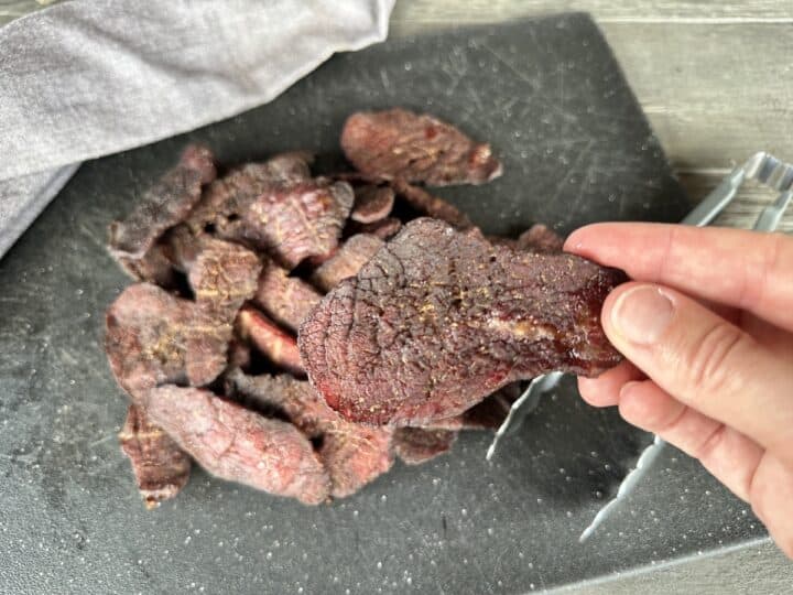 hand holding finished piece smoked beef jerky over a black cutting board with a pile of jerky