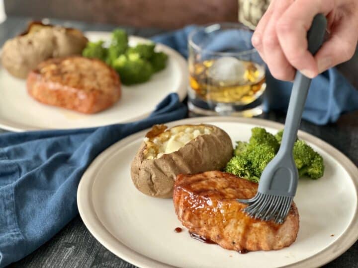 bourbon glazed pork chop being brushed with glaze on white plate with baked potato broccoli and glass of bourbon