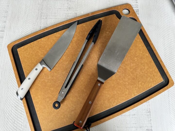 brown cutting board with chefs knife tongs and large stainless steel spatula