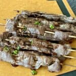piled up teriyaki beef skewers on cutting board garnished with green onions and sesame seeds
