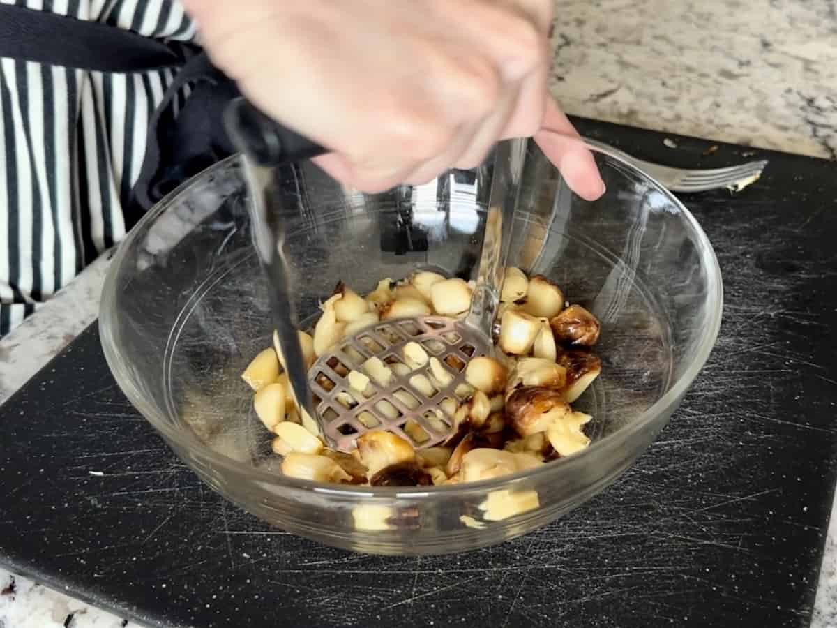 glass bowl of smoked garlic cloves being mashed with potato masher