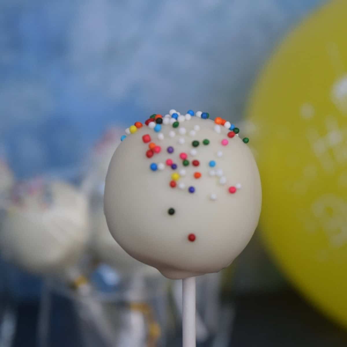 white cake pop with rainbow sprinkles and more cake pops and a yellow balloon in the background