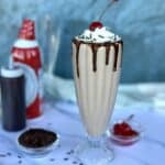 thick chocolate milkshake in classic milkshake glass whipped cream chocolate sauce sprinkles and cherries in background