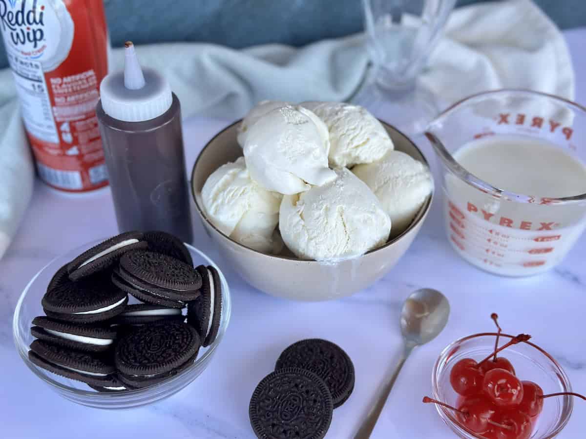 oreo milkshake ingredients chocolate syrup in plastic bottle scooped vanilla ice cream in brown bowl Oreo cookies in glass bowl canned whipped cream glass measuring cup of milk cherries