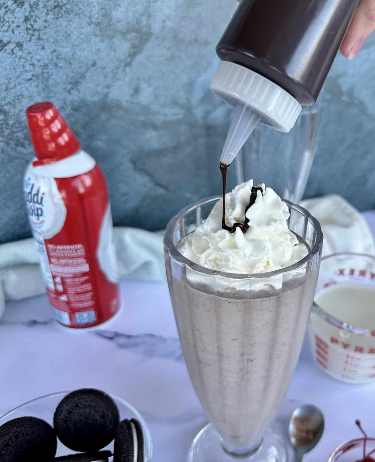 canned whipped cream next to prepared milkshake topped with whipped cream getting drizzle of chocolate syrup