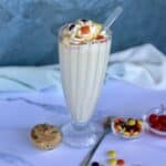 prepared peanut milkshake topped with whipped cream caramel sauce and candies white marbled background extra candies and cherries in glass bowls green kitchen towel