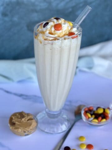 prepared peanut milkshake topped with whipped cream caramel sauce and candies white marbled background extra candies and cherries in glass bowls green kitchen towel