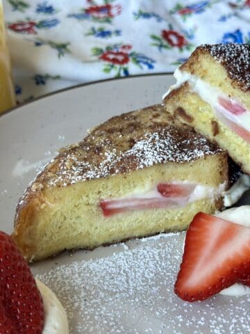 cheesecake French toast with strawberries on tan plate glass of orange juice and flower apron on white table