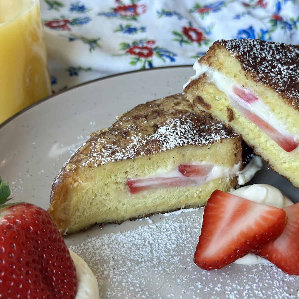 cheesecake French toast with strawberries on tan plate glass of orange juice and flower apron on white table