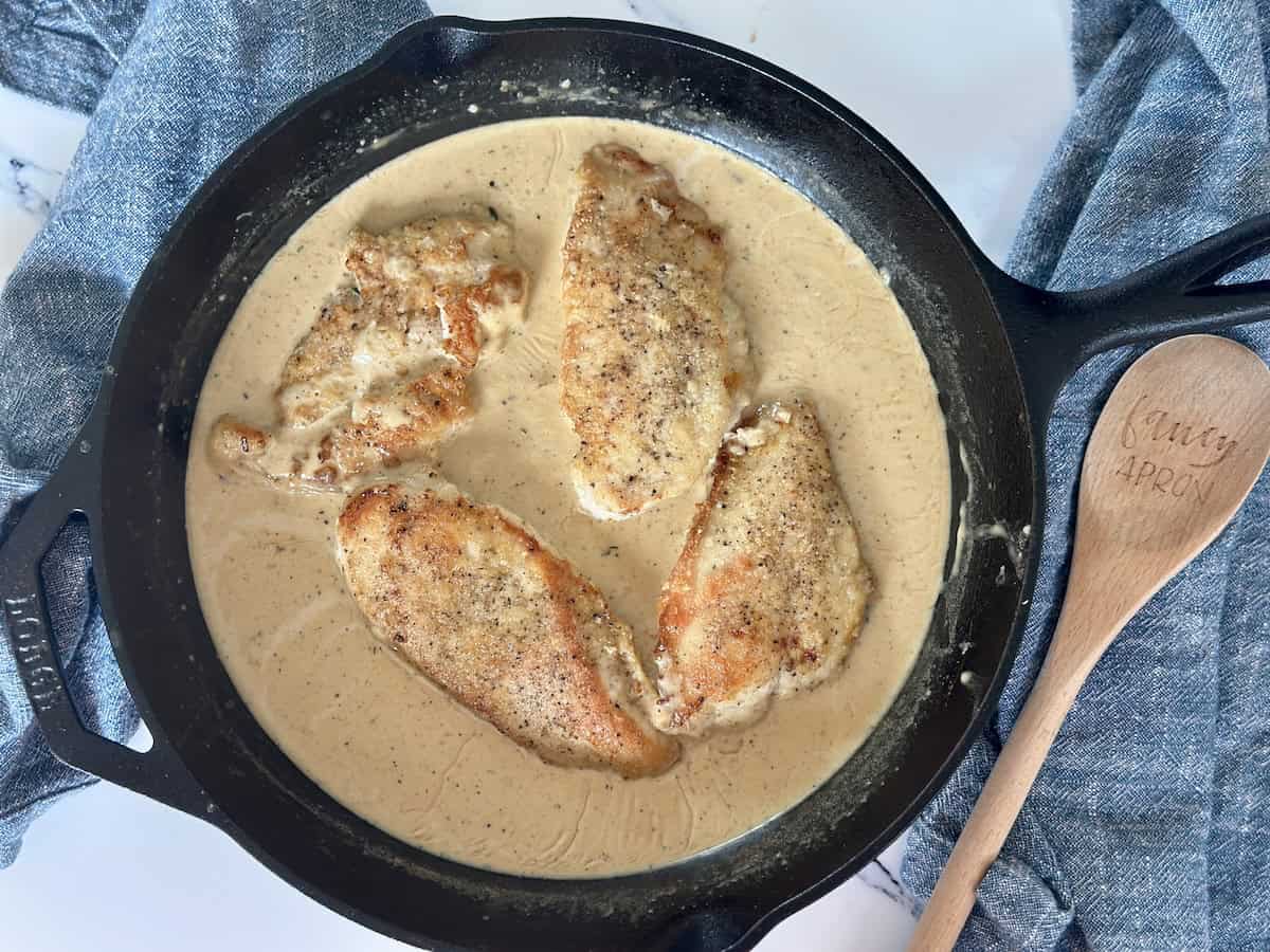 four chicken breasts cooked in black cast iron pan with cream sauce blue linen napkins wood spoon