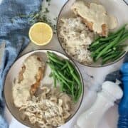 breaded chicken breast on tan plate with cream sauce bow tie pasta green beans blue napkin half of a lemon sprigs of thyme fork and knife white salt grinder