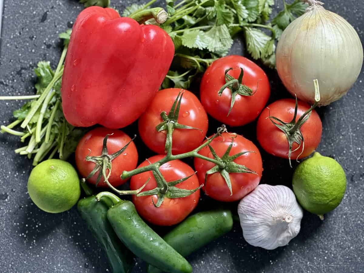 red bell pepper tomatoes on the vine yellow onion head of garlic lime jalapeño pepper pile of cilantro on black cutting board