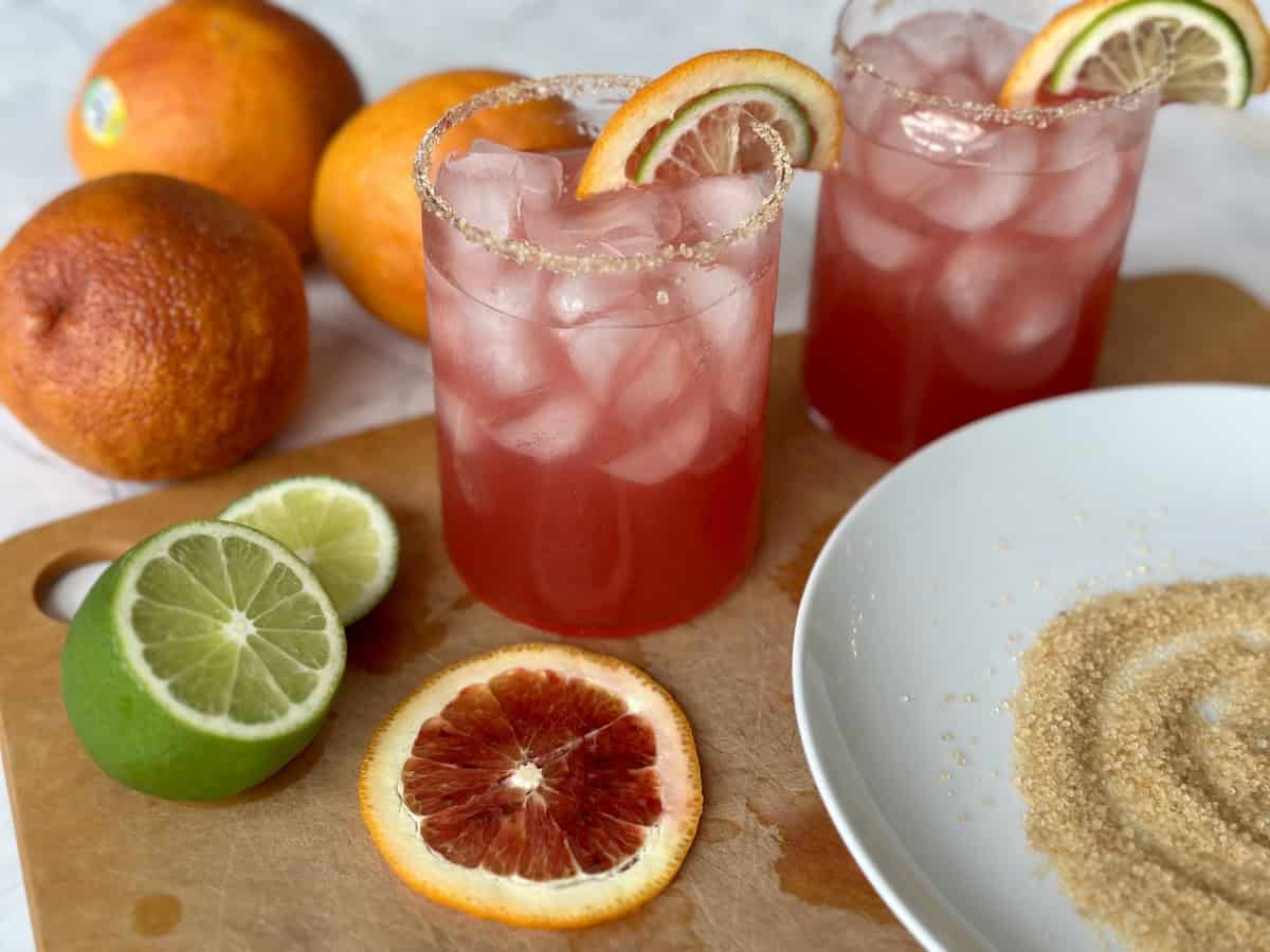 2 blood orange margaritas on wood cutting board white plate with turbinado sugar slices of lime and blood oranges