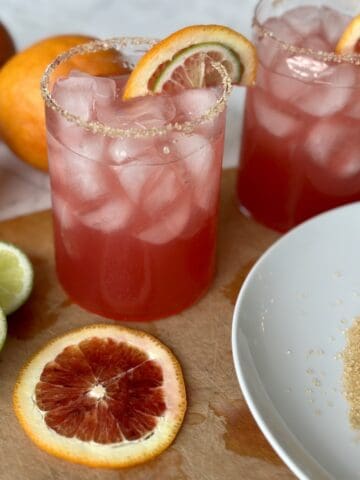 2 blood orange margaritas on wood cutting board white plate with turbinado sugar slices of lime and blood oranges
