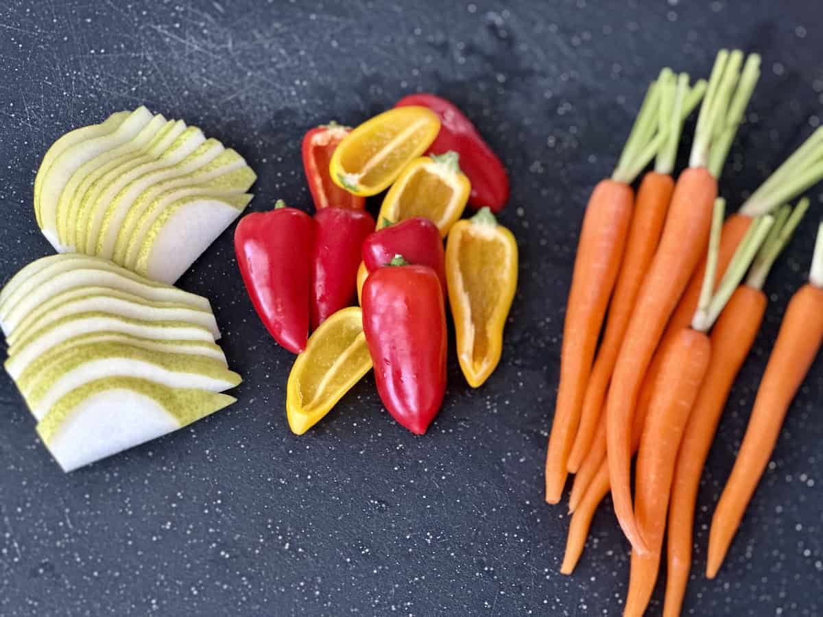 sliced pear mini bell pepper and thin carrots with the tops on black cutting board