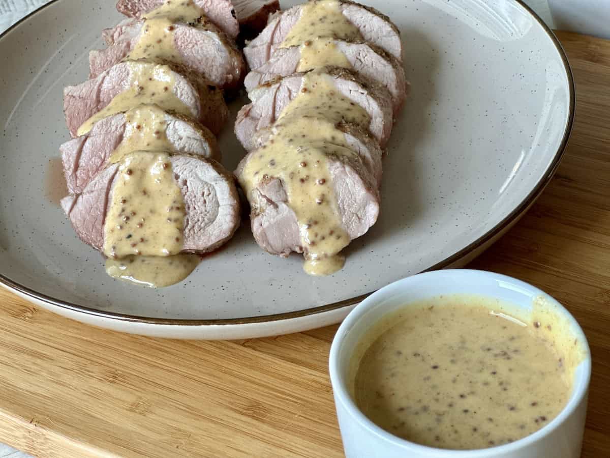 sliced pork tenderloin on light brown plate white cup of whole grain honey mustard sauce all on wood cutting board