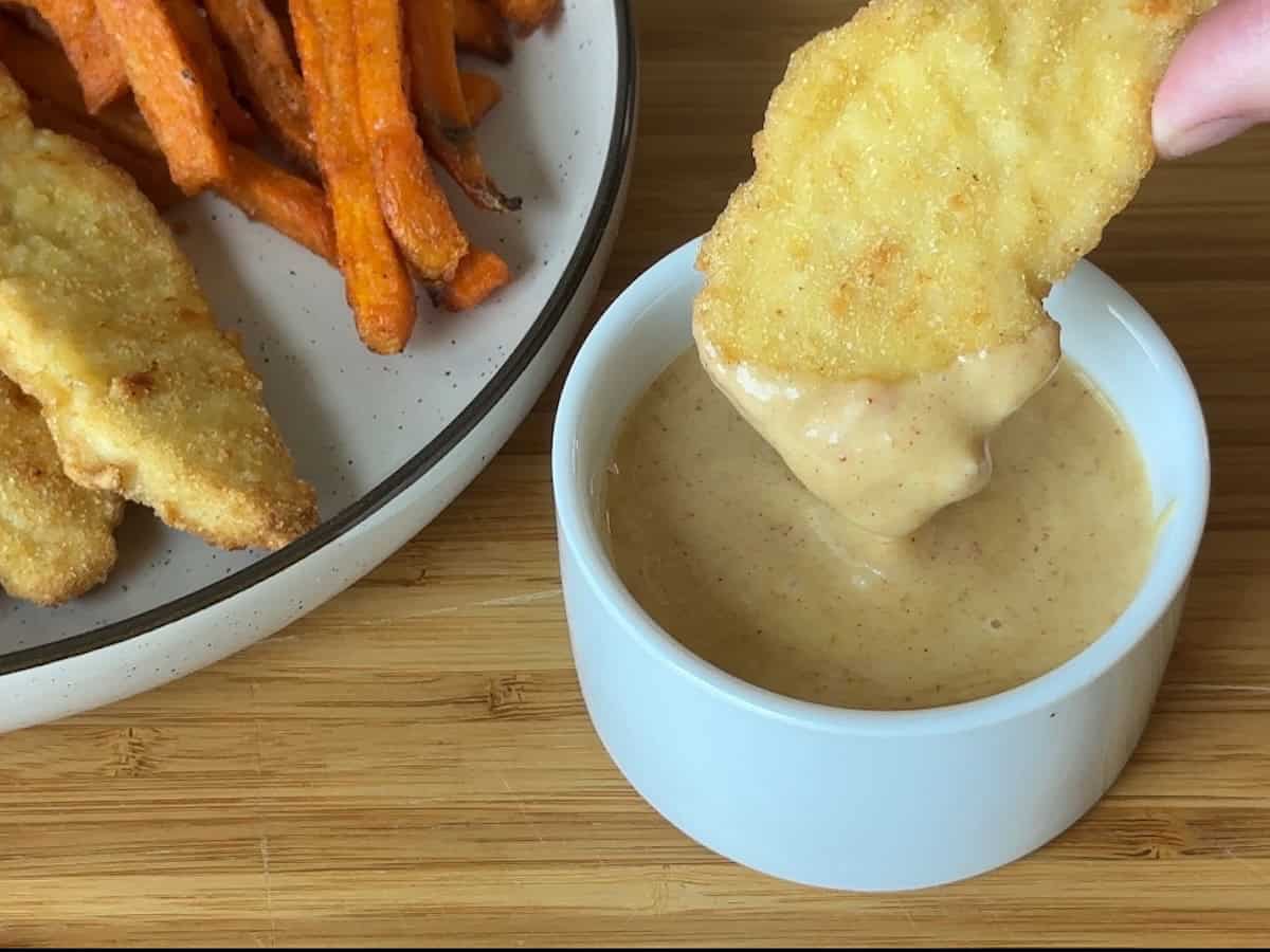 chicken tender dipped in honey mustard sauce in small white bowl next to plate of chicken tenders and sweet potato fries