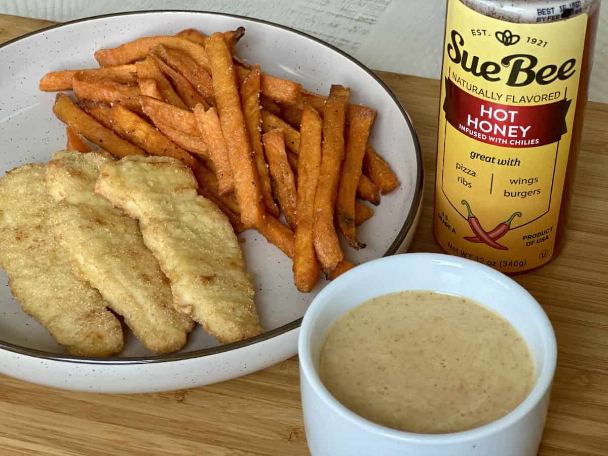 tan plate with chicken tenders and sweet potato fries next to white bowl of sauce and bottled hot honey