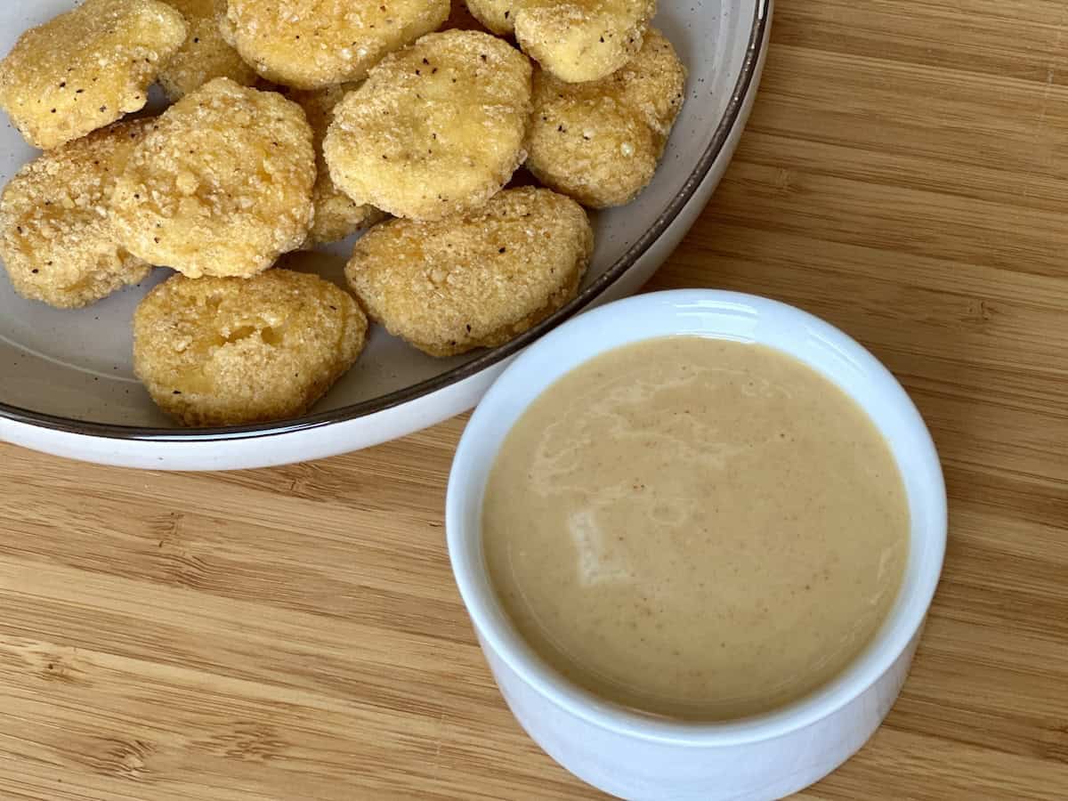 white bowl of honey mustard sauce next to tan plate of chicken nuggets on wood cutting board