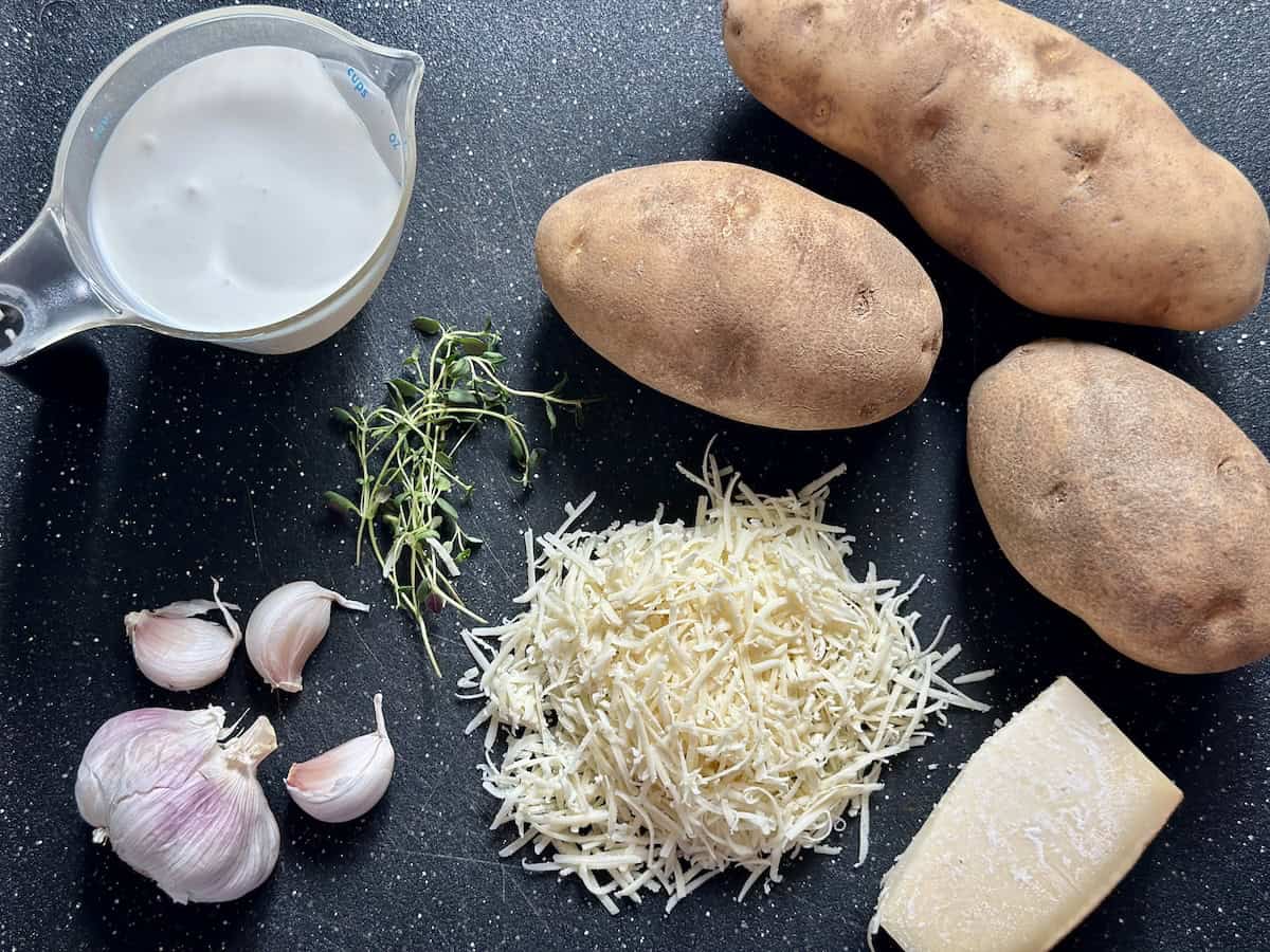 russet potatoes cup of heavy cream head of garlic sprigs of thyme grated gruyere cheese on black cutting board