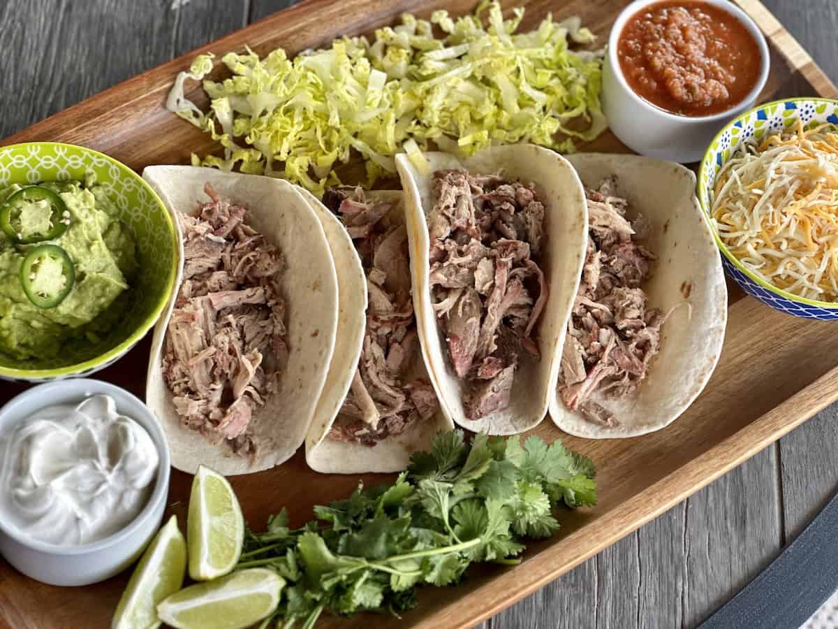 4 pulled pork soft tacos on wood board with bowls of sour cream guacamole shredded cheese Sala with lettuce cilantro and lime