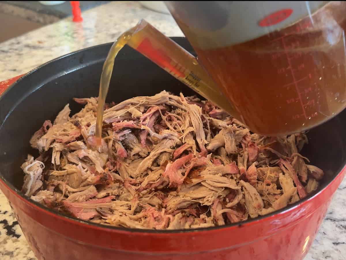 juices from fat separator pouring over red dutch oven of pulled pork