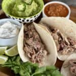 two soft tacos with shredded pork green bowl of guacamole white bowl sour cream white bowl salsa cilantro garnish on wood board