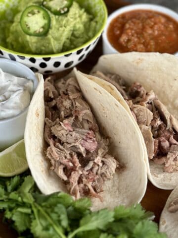 two soft tacos with shredded pork green bowl of guacamole white bowl sour cream white bowl salsa cilantro garnish on wood board