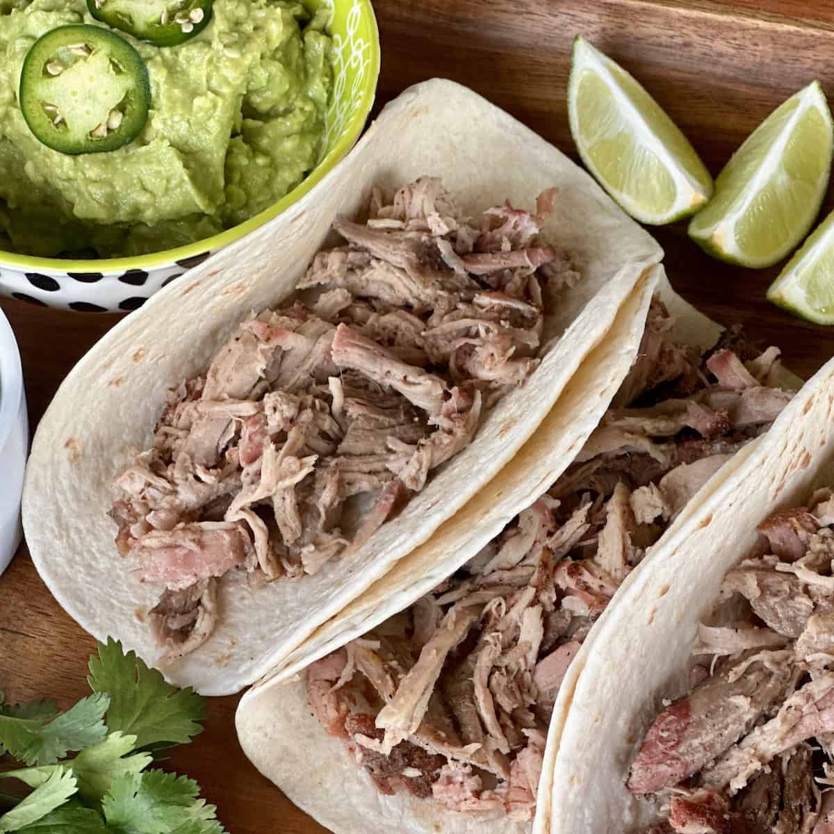 3 soft tacos filled with shredded pork on wood board with bowl of guacamole lime wedges and cilantro garnish