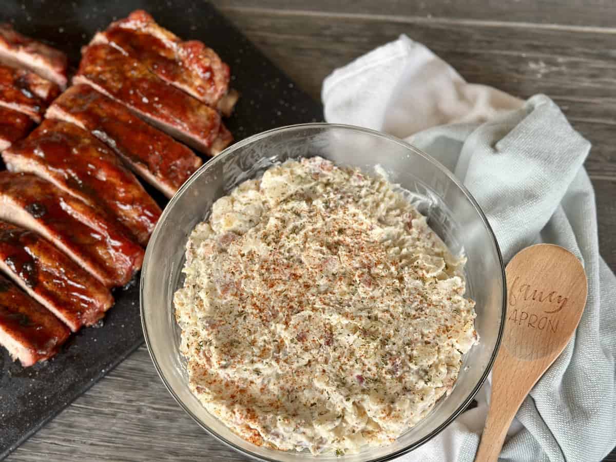 A glass bowl filled with potato salad and a wooden spoon with sliced ribs on cutting board