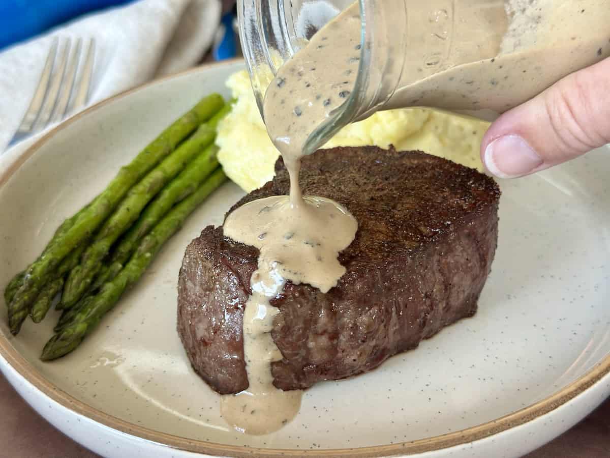 pouring peppercorn cream sauce over cooked steak on plate with mashed potatoes and asparagus