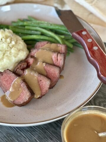 sliced steak with miso sauce on plate with green beans mashed potatoes and steak knife