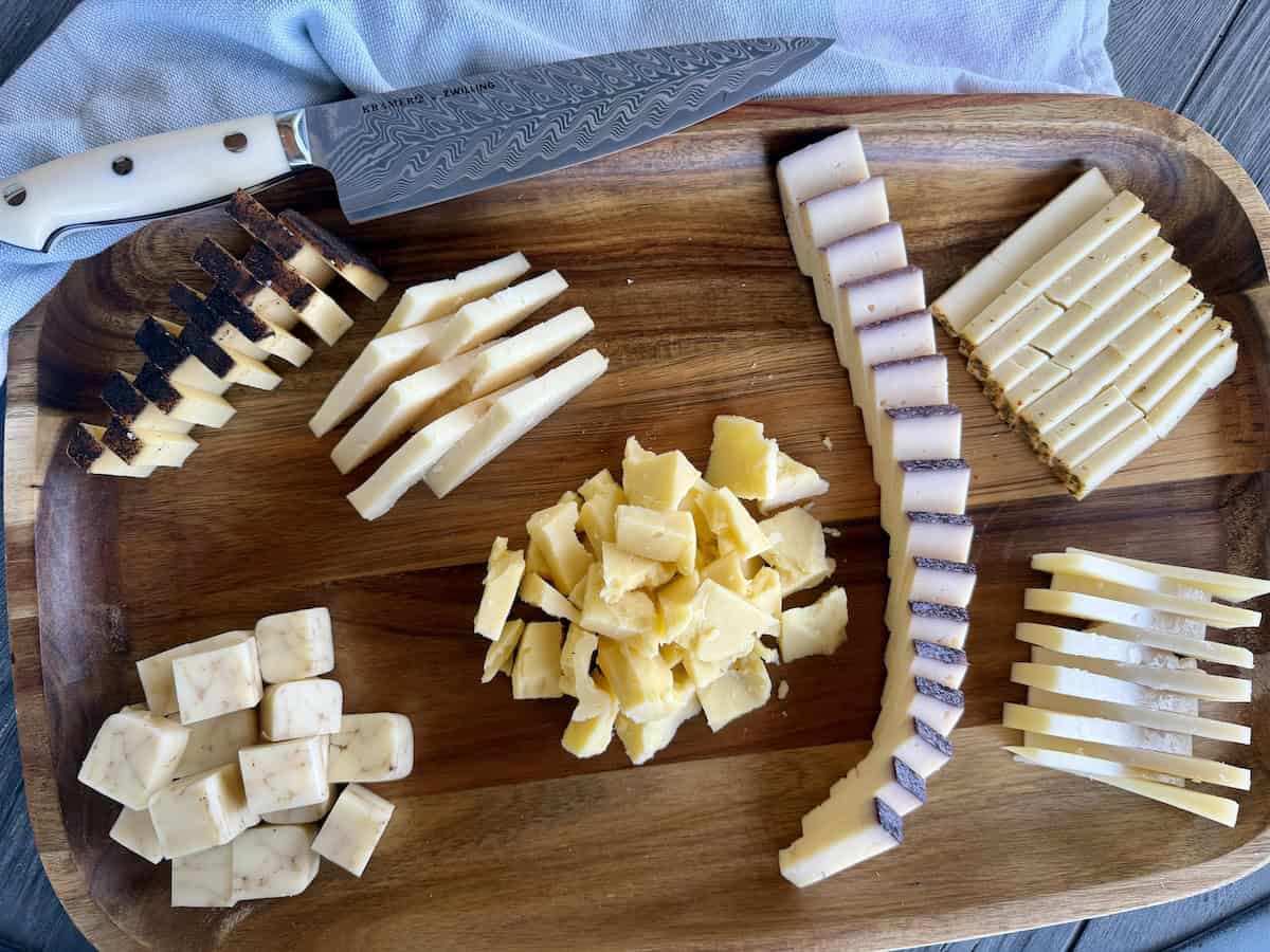 cut hard cheeses styled differently on wood charcuterie board chef's knife and green kitchen towel