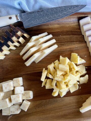 cut hard cheeses styled differently on wood charcuterie board chef's knife and green kitchen towel