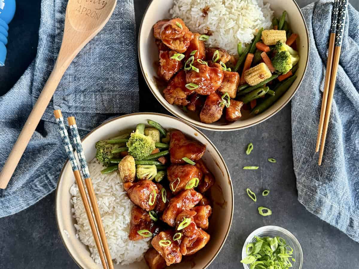 two bowls bourbon chicken with rive stir fried vegetables chopsticks blue napkins