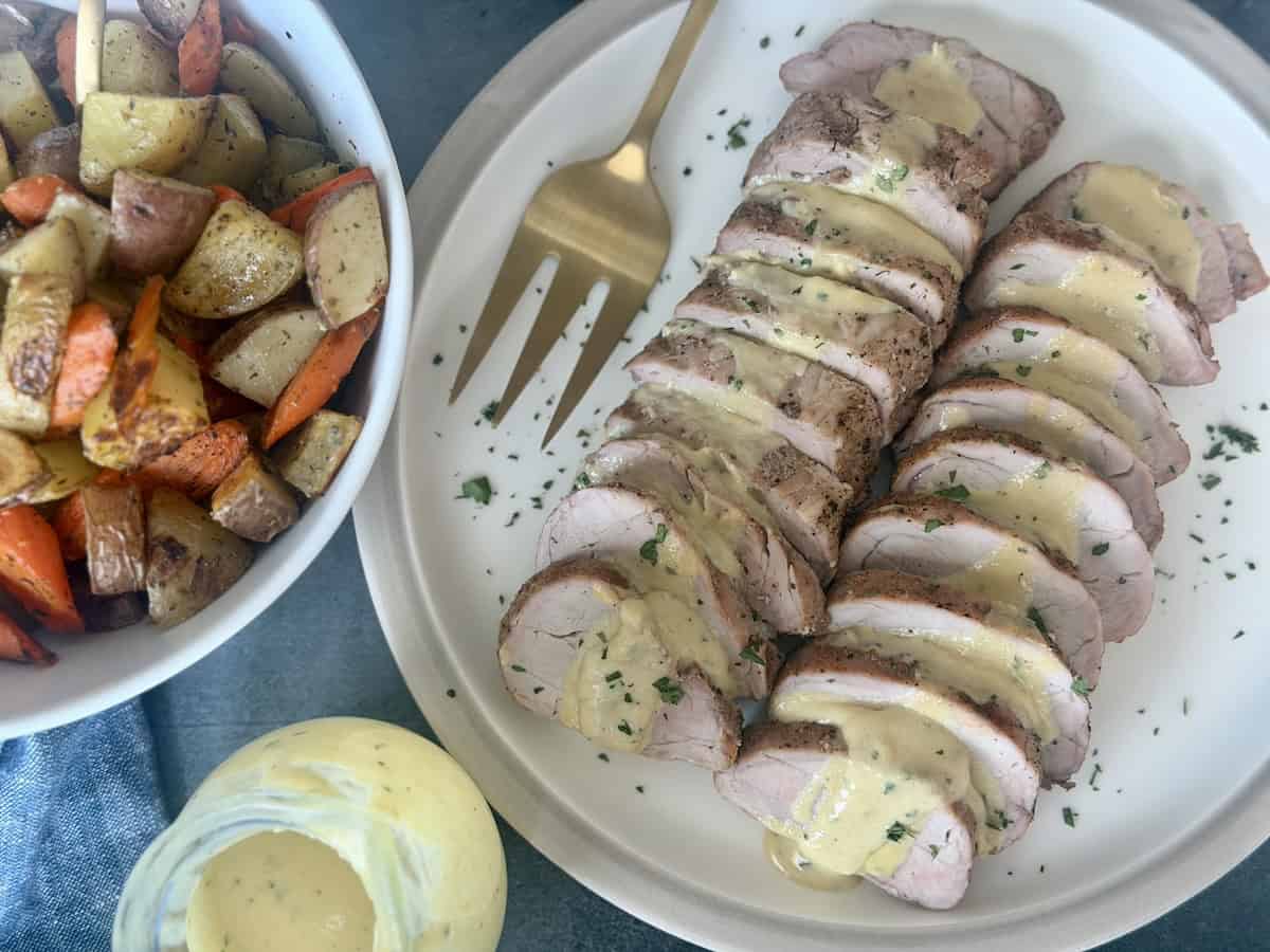 sliced pork tenderloin creamy mustard sauce on white plate roasted gold fork blue napkin vegetables 