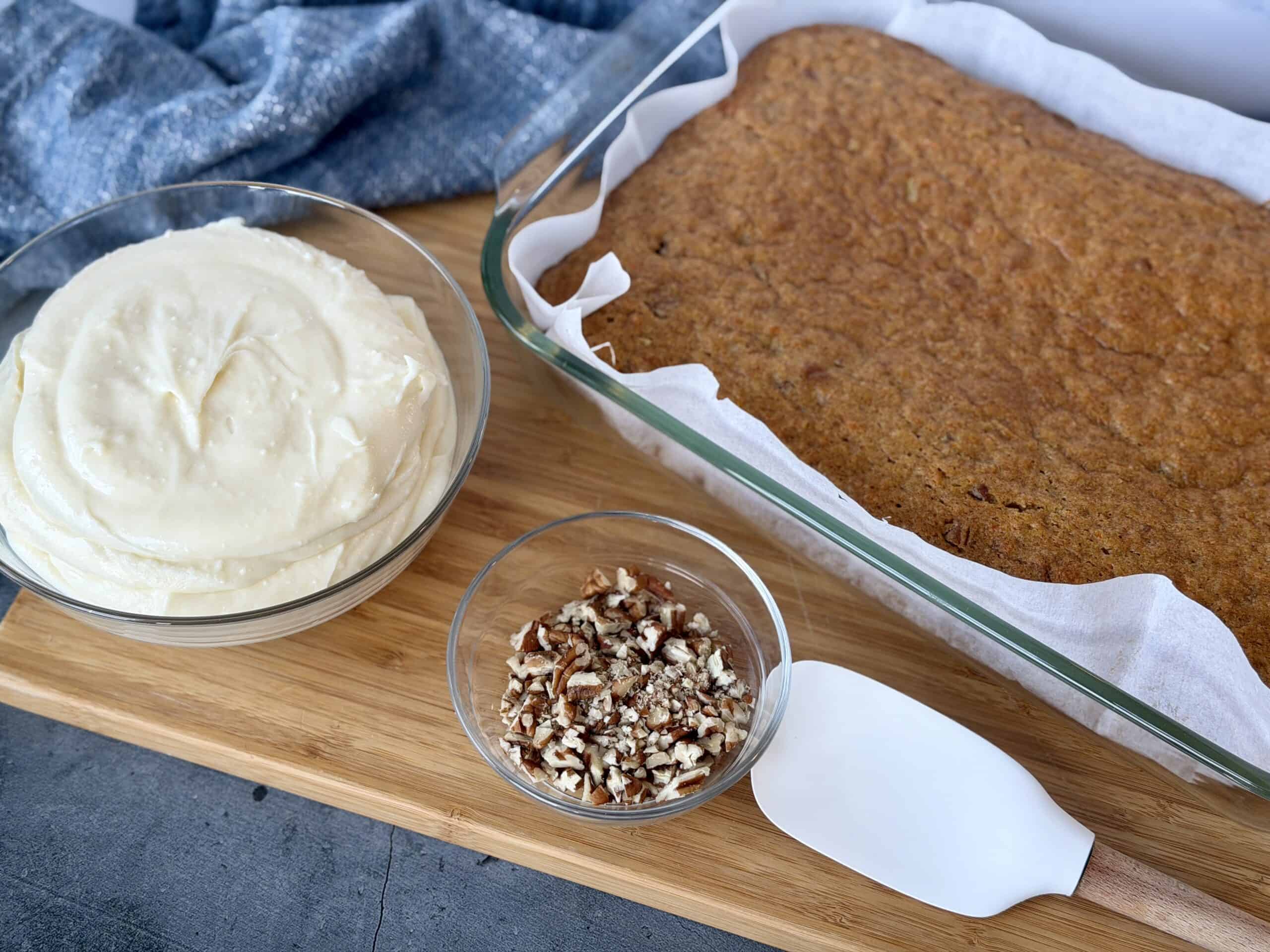 baked carrot cake in glass dish bowl of cream cheese frosting bowl of chopped pecans white spatula wood cutting board blue cloth napkin