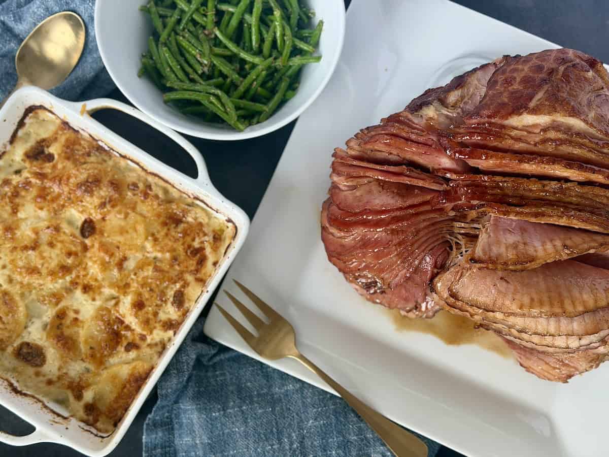 cooked spiral ham on white plate white bowl of green beans baking dish of au gratin potatoes 